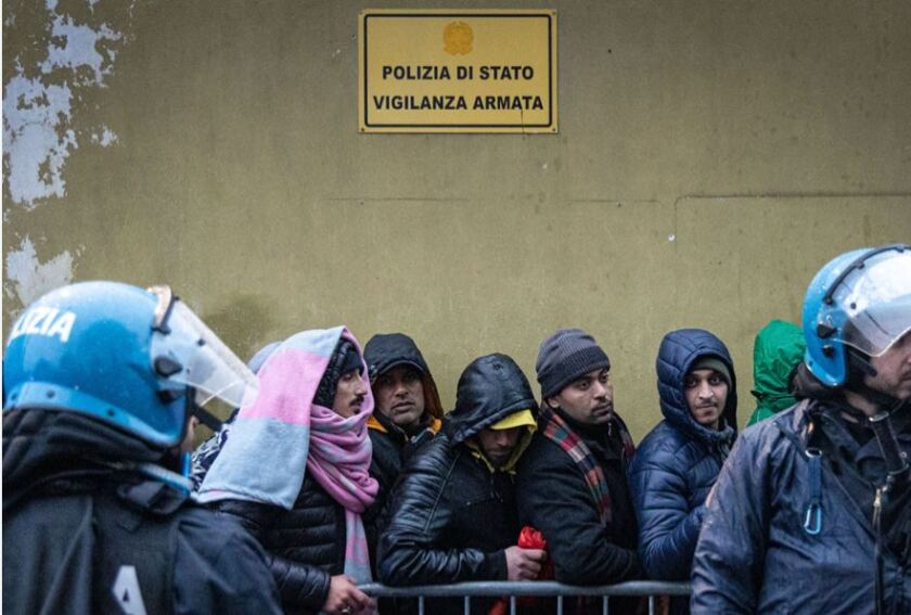 stranieri in fila davanti ad un muro con la scritta "polizia di stato, vigilanza armata"